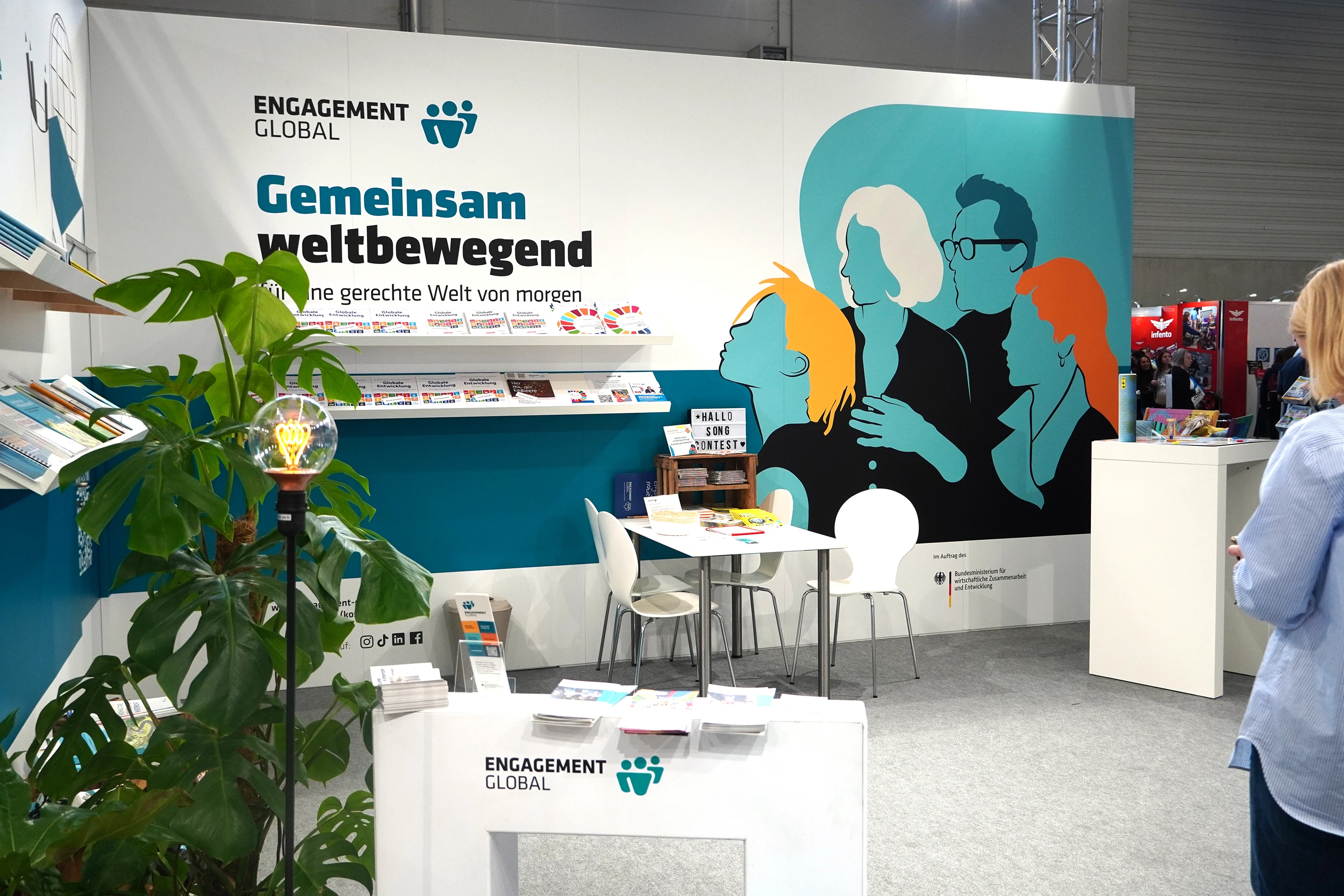 Messestand von Engagement Global mit der Wandgrafik „Gemeinsam weltbewegend“, Broschürenregalen und Beratungstisch