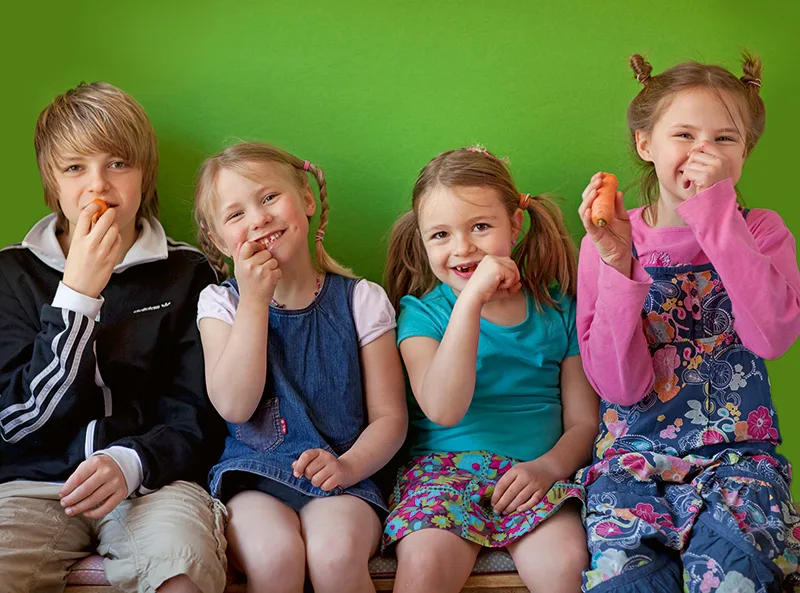 Vier Kinder essen lachend Möhren vor grüner Wand